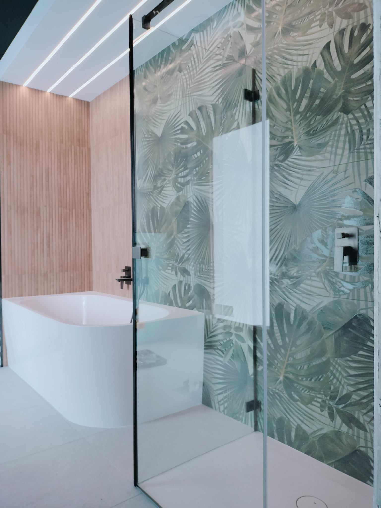 Nowoczesna łazienka z kabiną prysznicową ze szkła hartowanego, wolnostojącą białą wanną i ścianą wyłożoną płytkami w tropikalne liście, oświetlenie sufitowe LED.