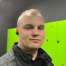 Klima&Systemy Radosław Malinowski - Mężczyzna w czarnej kurtce na tle jaskrawozielonych szafek w szatni z czarnym sufitem i widocznym głośnikiem.