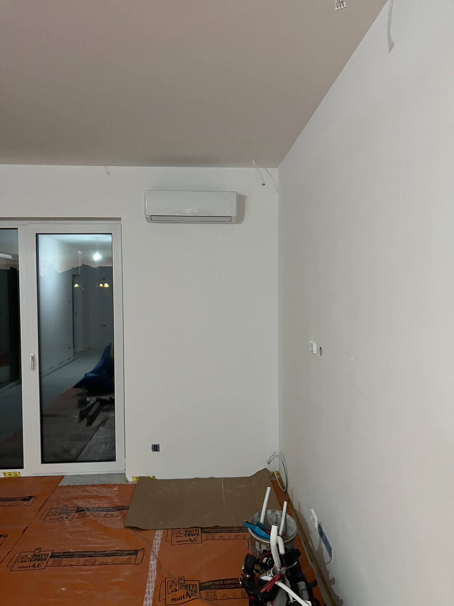 Biały klimatyzator na ścianie w jasnym pomieszczeniu, widoczne zabezpieczenia podłogi i narzędzia instalacyjne w rogu, okno balkonowe z widokiem na sąsiednie pomieszczenie.