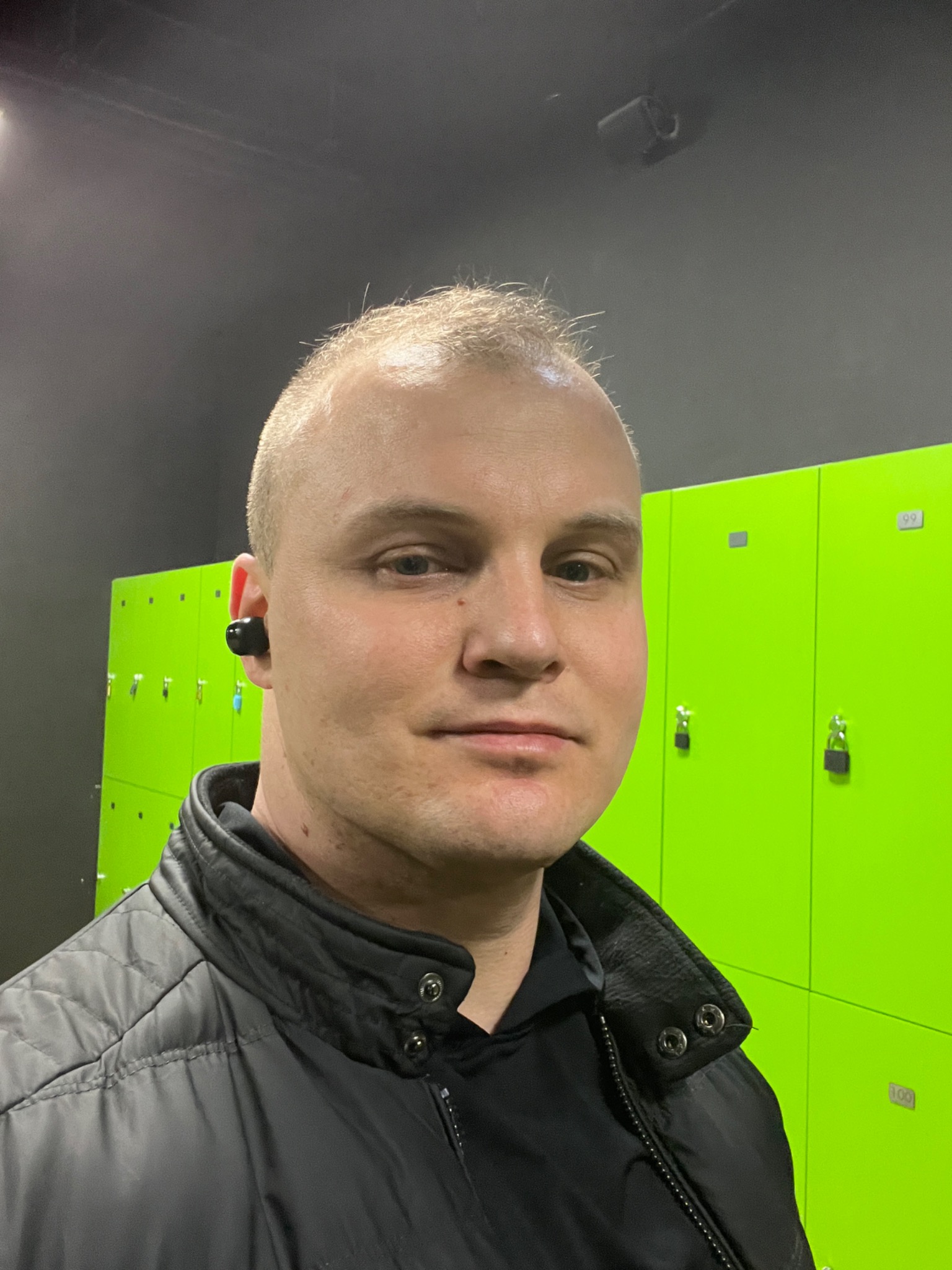 Mężczyzna w czarnej kurtce na tle jaskrawozielonych szafek w szatni z czarnym sufitem i widocznym głośnikiem.