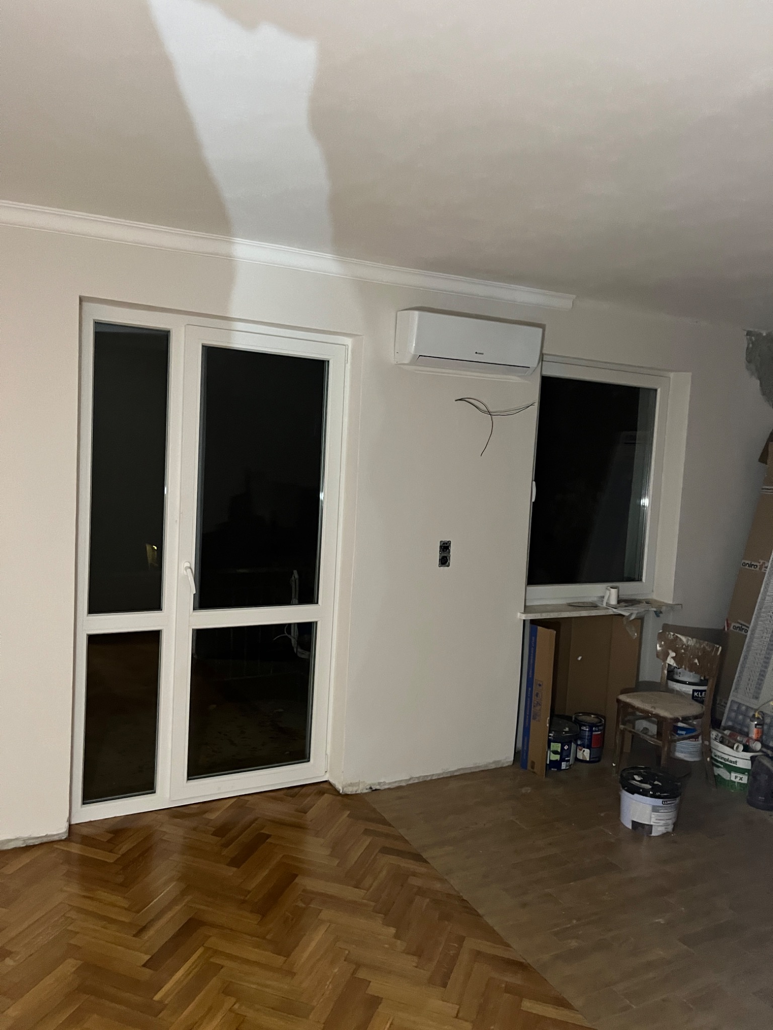 Biała jednostka klimatyzacji zamontowana nad oknem w pokoju z parkietem w jodełkę, widoczne kable, puszka elektryczna, okno balkonowe i puszki z farbą w tle.