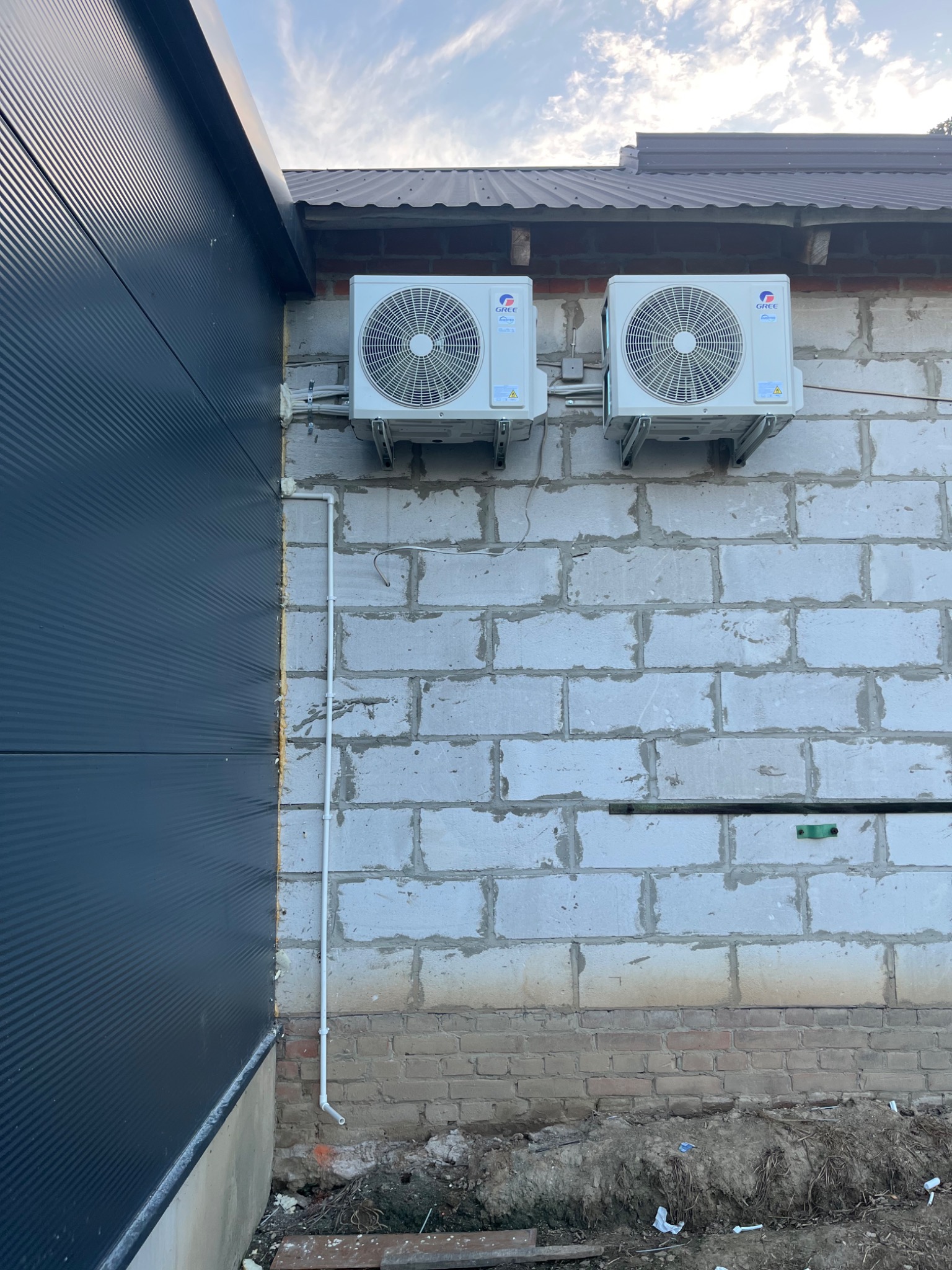 Dwa białe klimatyzatory marki Gree zamontowane na ścianie z białych pustaków, widoczne przewody instalacyjne i fragment elewacji budynku z blachy trapezowej w kolorze grafitowym.