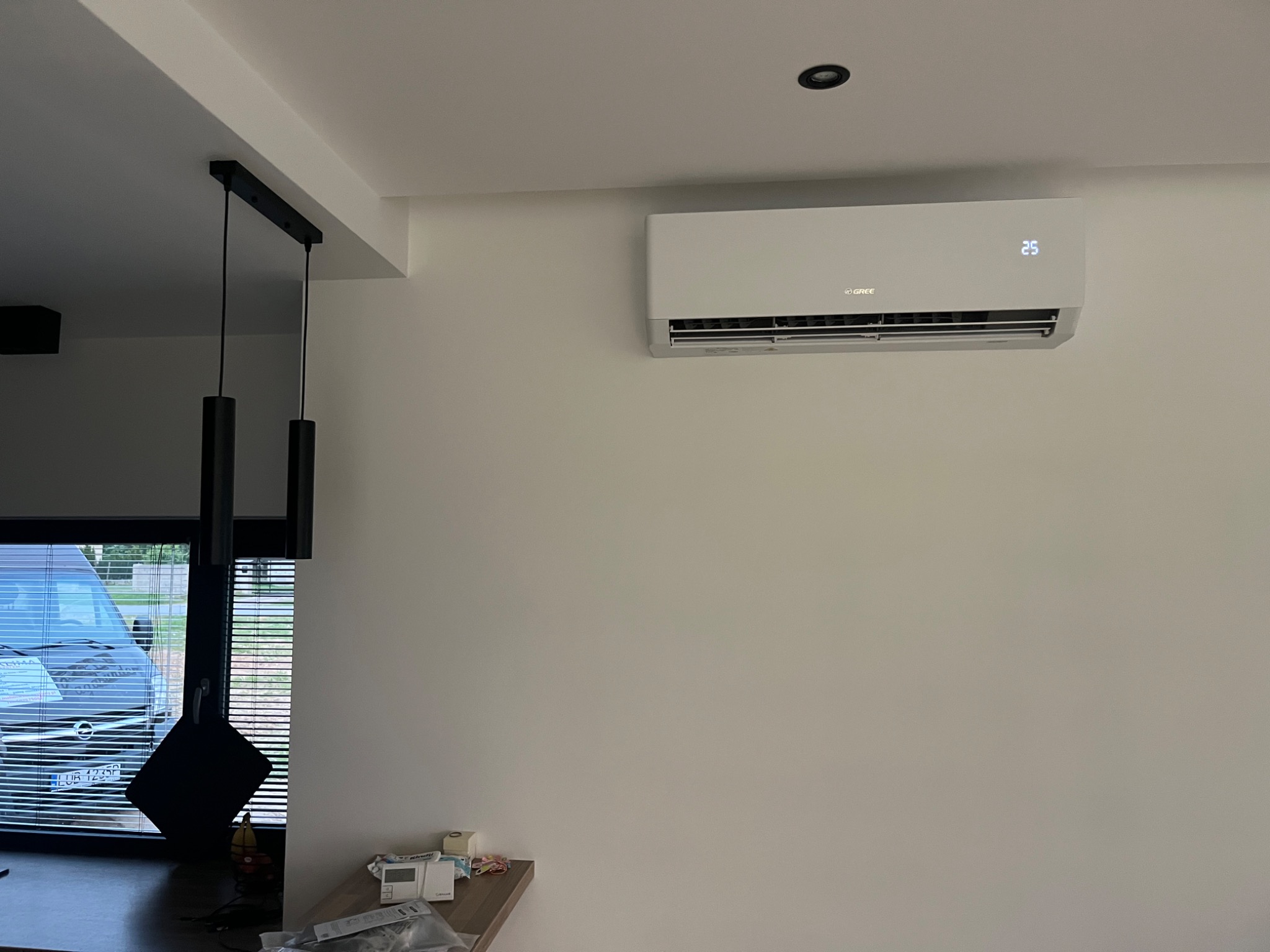 Biała klimatyzacja Gree zamontowana na jasnej ścianie w nowoczesnym wnętrzu, widoczna temperatura 25 stopni Celsjusza, obok okno z żaluzjami i fragment czarnej lampy wiszącej.