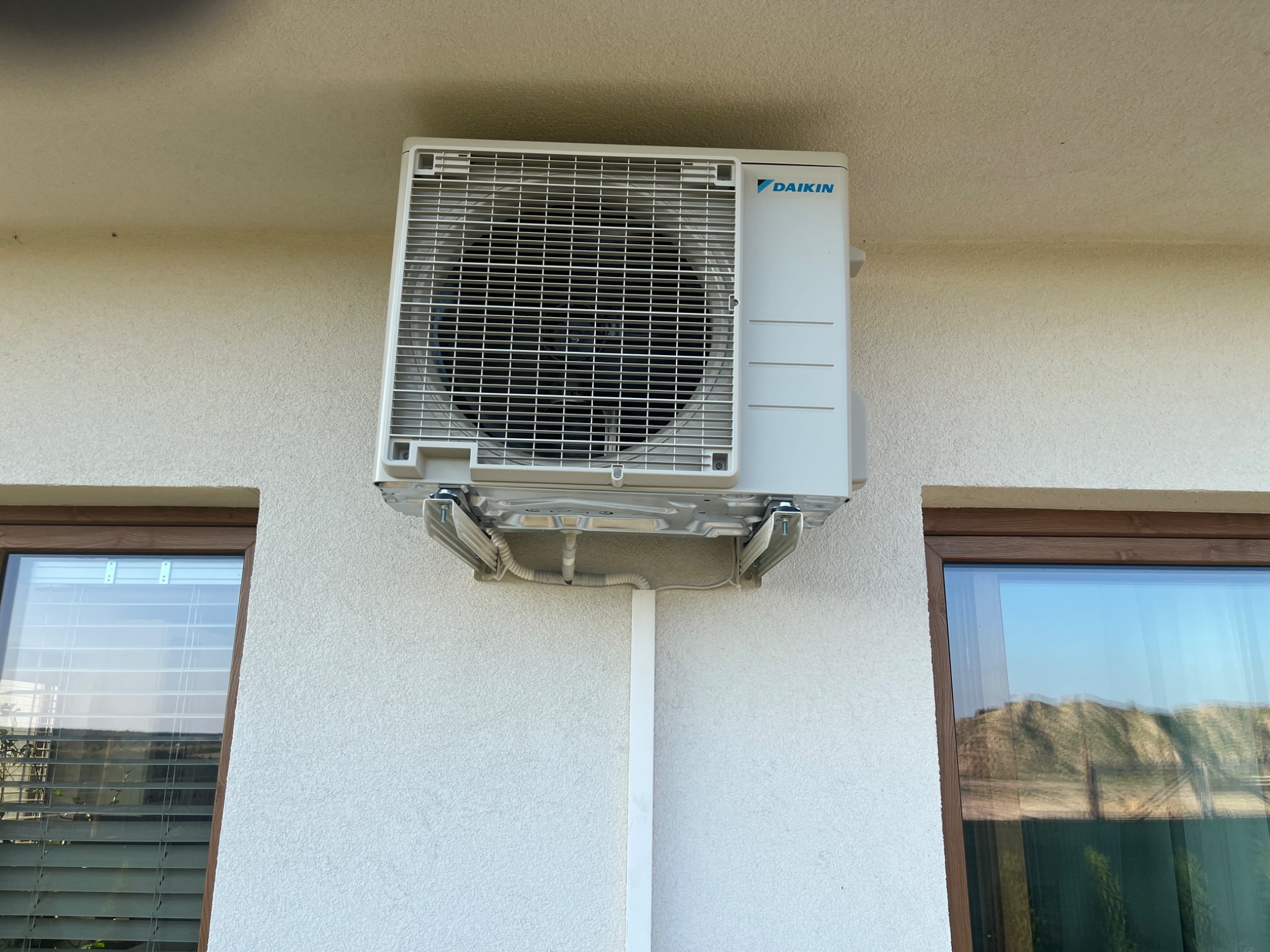 Zewnętrzna jednostka klimatyzacji Daikin zamontowana na elewacji budynku, z widocznymi oknami po bokach i pionową listwą maskującą instalację elektryczną.