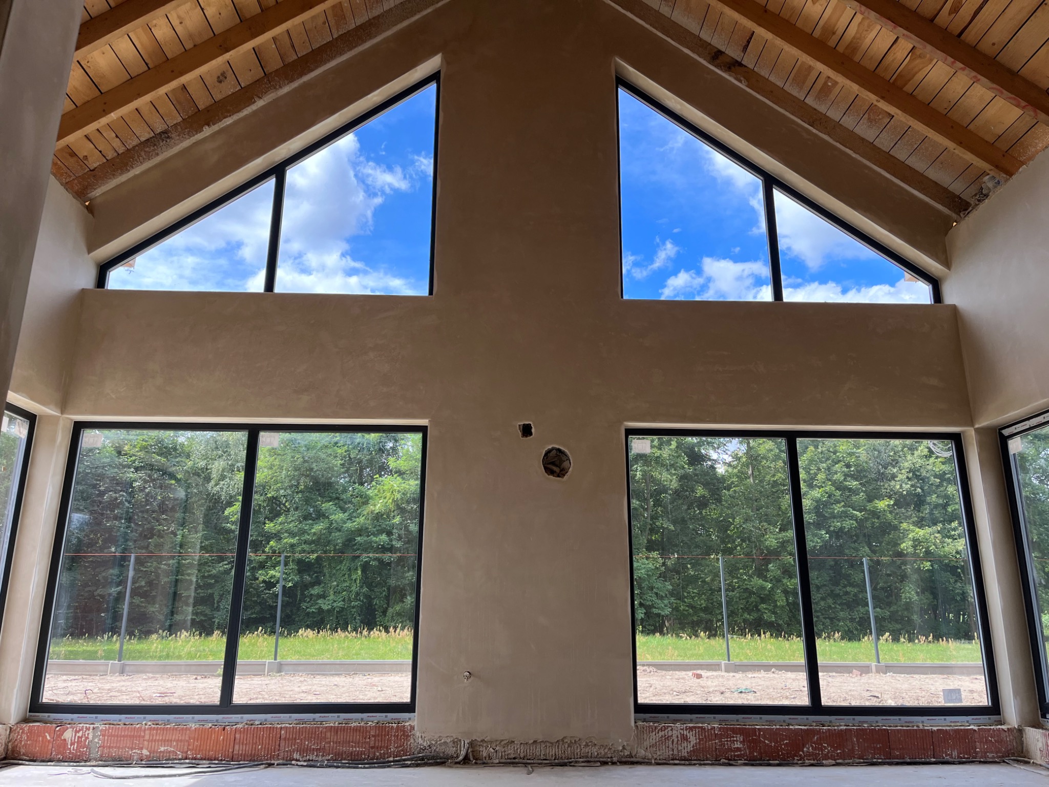 Wnętrze w stanie surowym z nałożonymi tynkami i dużymi oknami z widokiem na zieleń, drewniany strop, widoczne instalacje elektryczne w ścianie.