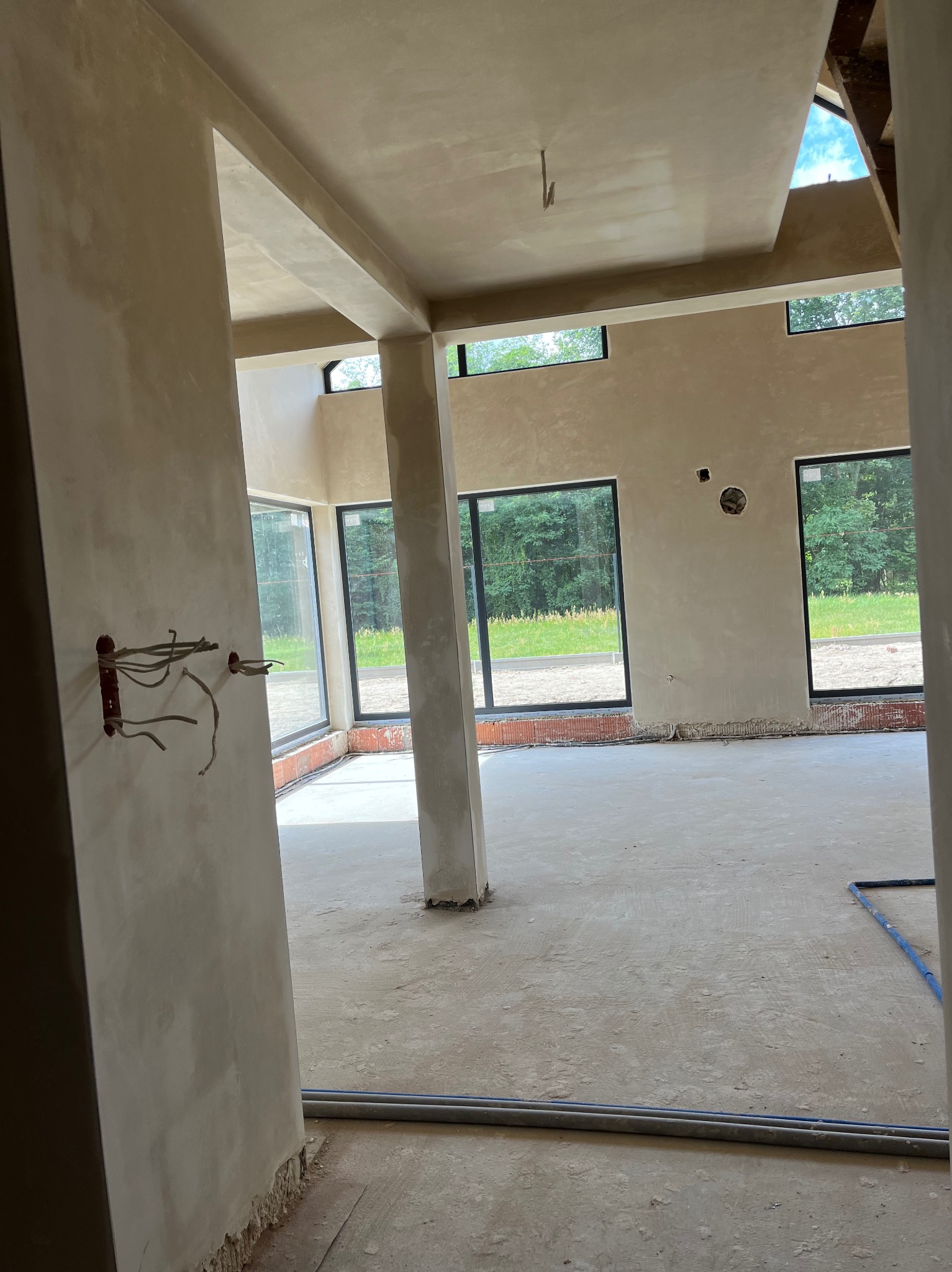 Wnętrze budynku w stanie surowym po nałożeniu jasnej, gładkiej warstwy tynku na ściany i sufit, widoczne wystające przewody elektryczne i rury na podłodze, duże okna z widokiem na zieleń na zewnątrz.