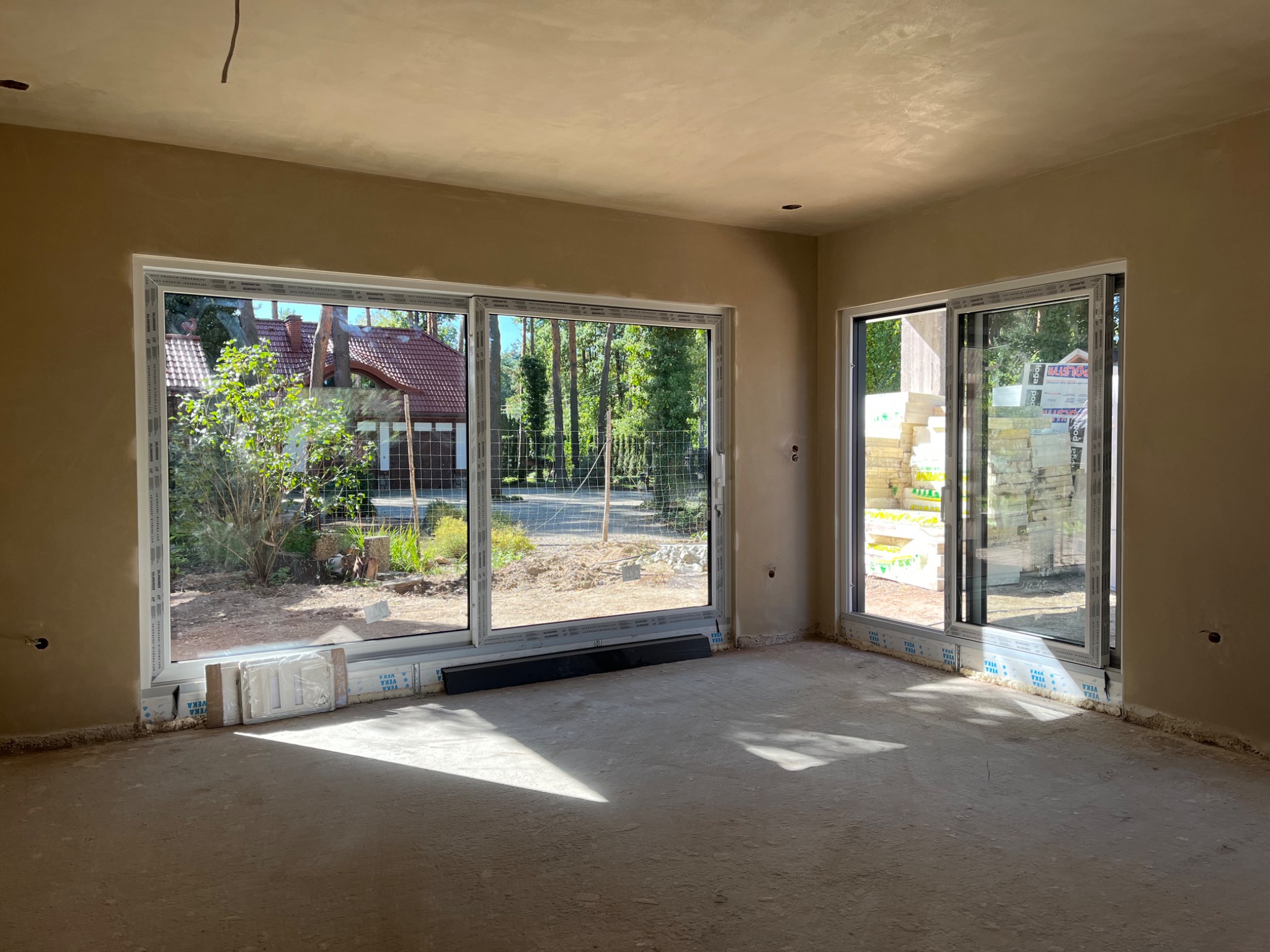 Wnętrze domu po nałożeniu gładzi gipsowej na ściany i sufit, widoczne duże okna przesuwne z widokiem na zewnątrz, na podłodze pozostałości po pracach wykończeniowych oraz puszki z osprzętem...