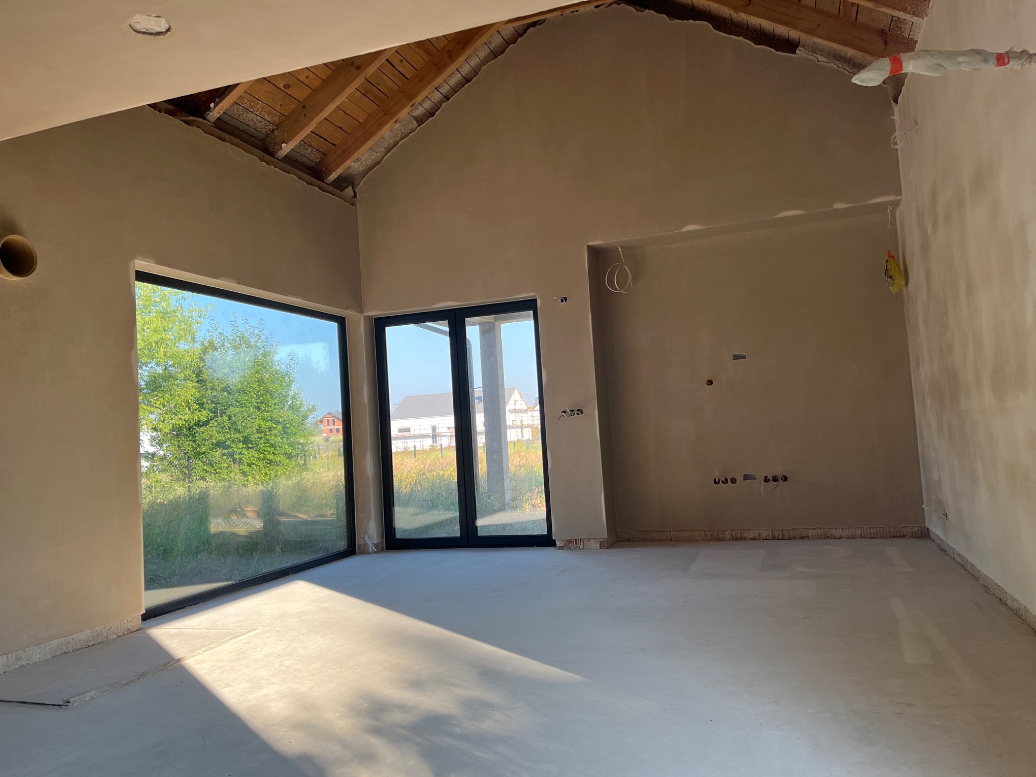 Wnętrze surowego domu po nałożeniu tynków, z widocznymi dużymi oknami i przygotowanym poddaszem, wpadające światło słoneczne podkreśla gładką powierzchnię ścian i podłogi.