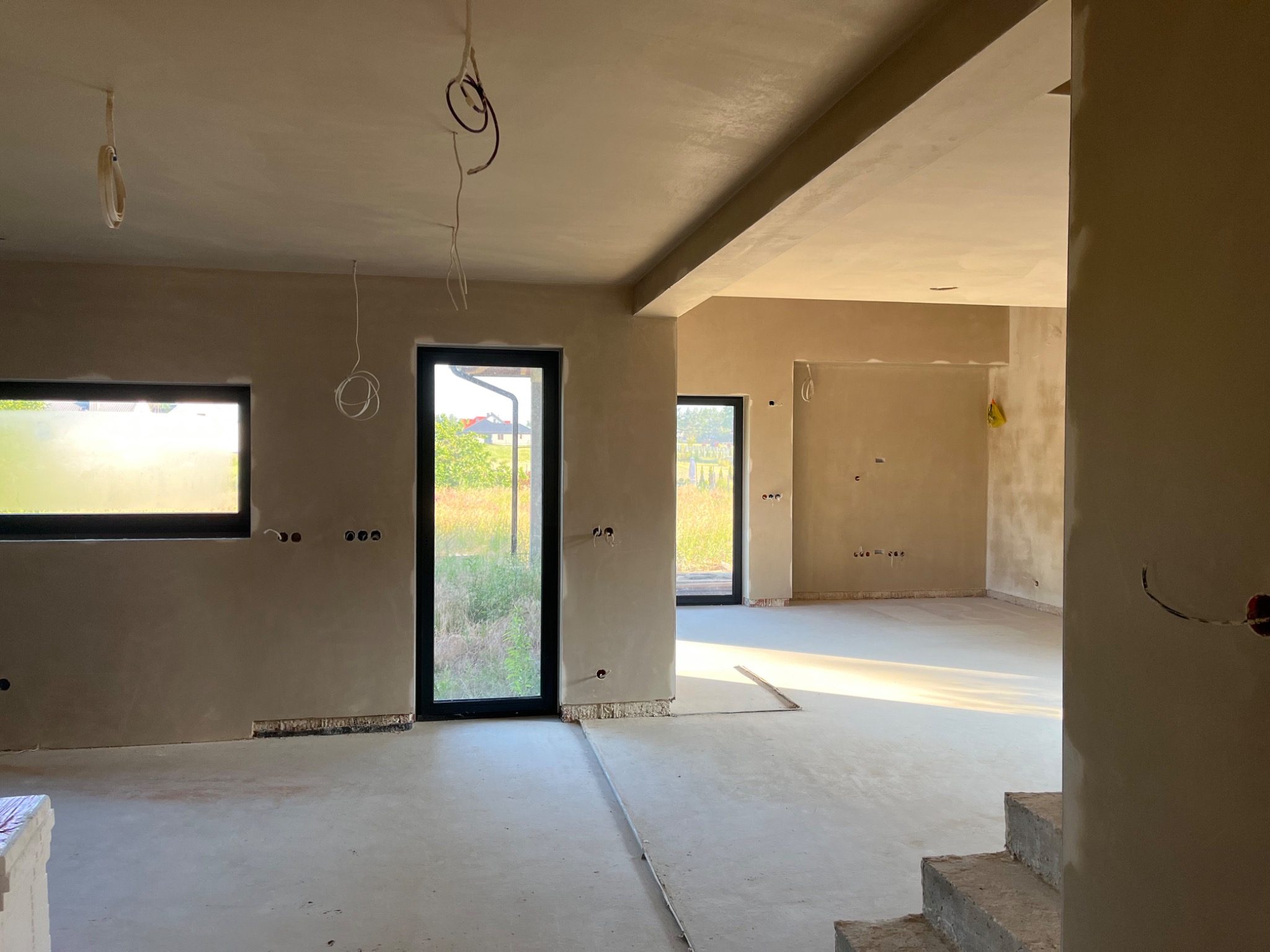 Wnętrze domu w trakcie prac wykończeniowych: świeżo otynkowane ściany, widoczne przewody elektryczne i fragment schodów w surowym stanie.