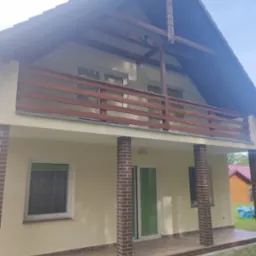 Elewacja domu jednorodzinnego z beżową farbą, drewnianą balustradą na balkonie i kolumnami z cegły klinkierowej na tarasie, widok z dołu na błękitne niebo.