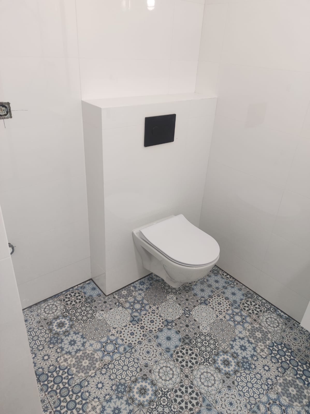 Nowoczesna, podwieszana toaleta z czarnym panelem sterującym na tle białych, błyszczących płytek ściennych i wzorzystej, niebiesko-szarej podłogi z płytek ceramicznych.