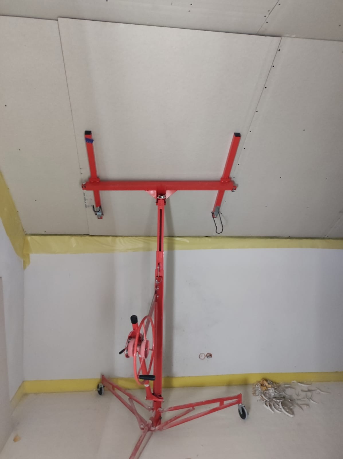 Montaż sufitu podwieszanego z płyt kartonowo-gipsowych przy użyciu windy do płyt, widoczne taśmy zabezpieczające na ścianach.