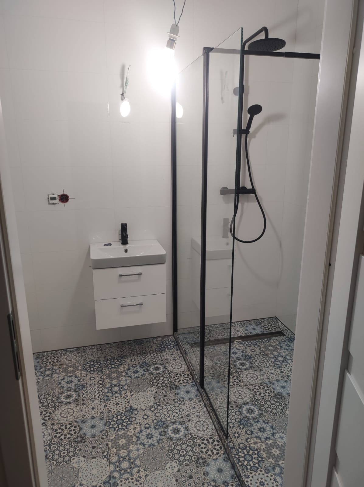 Nowoczesna łazienka z kabiną prysznicową typu walk-in z czarnymi ramami, białą umywalką z szafką i wzorzystą, patchworkową podłogą. W tle widoczne białe płytki ścienne i niedokończona instalacja...