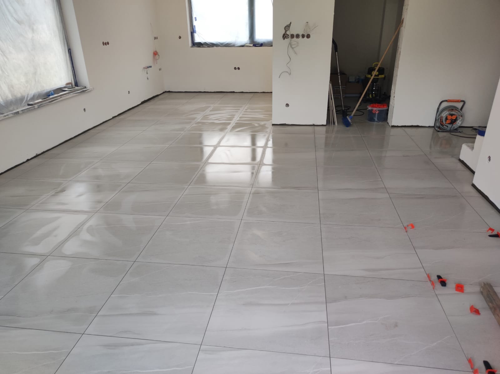 Jasnoszara, błyszcząca podłoga z płytek ceramicznych imitujących marmur, świeżo ułożona w pustym pomieszczeniu, z widocznymi plastikowymi krzyżykami dystansowymi i narzędziami w tle.