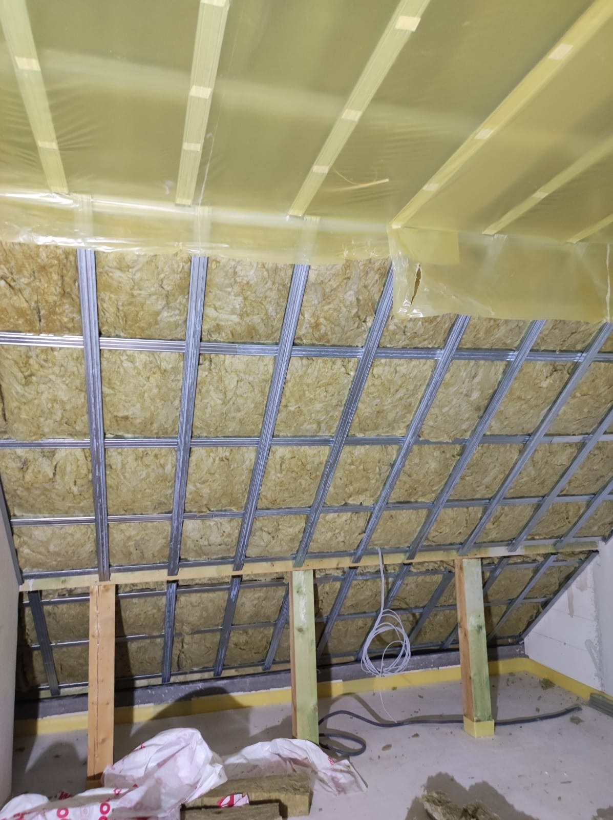 Montaż izolacji z wełny mineralnej w metalowym stelażu poddasza, zabezpieczonej folią paroizolacyjną, widoczne drewniane podpory i zwisające przewody elektryczne.