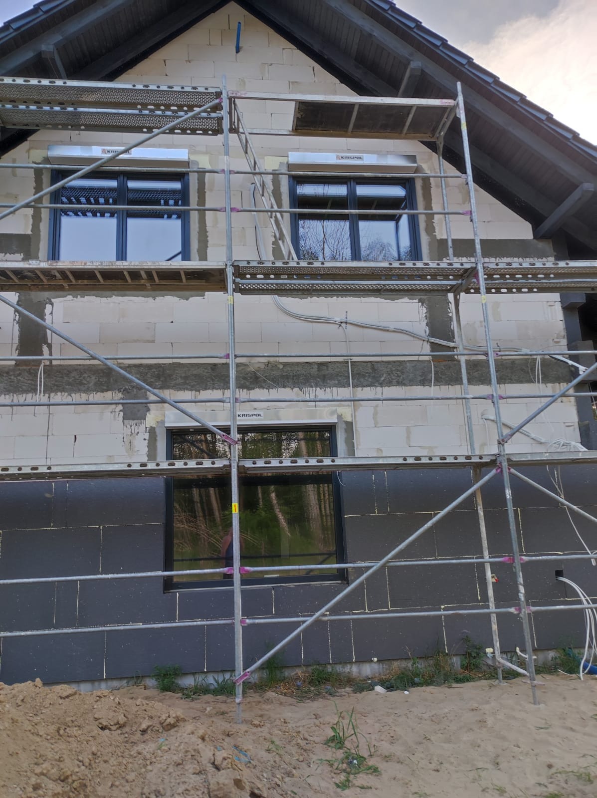 Budynek w trakcie ocieplania z widocznym rusztowaniem, czarną izolacją termiczną na dolnej części elewacji oraz szarymi bloczkami na górze, okna z zamontowanymi roletami zewnętrznymi.
