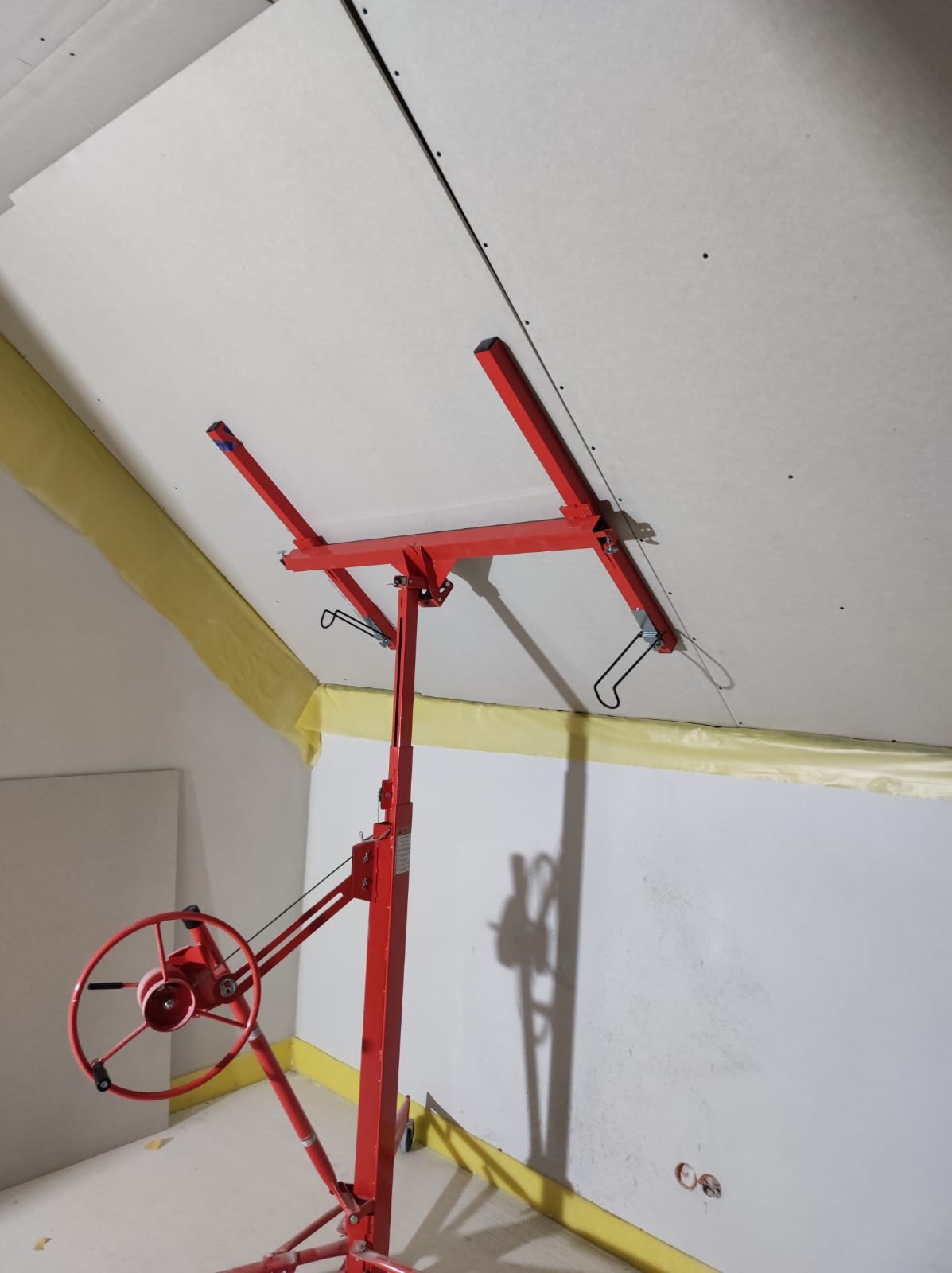 Czerwony podnośnik do płyt gipsowo-kartonowych podtrzymuje płytę przy skośnym suficie poddasza, widoczne łączenia płyt i żółta taśma zabezpieczająca przy ścianach.