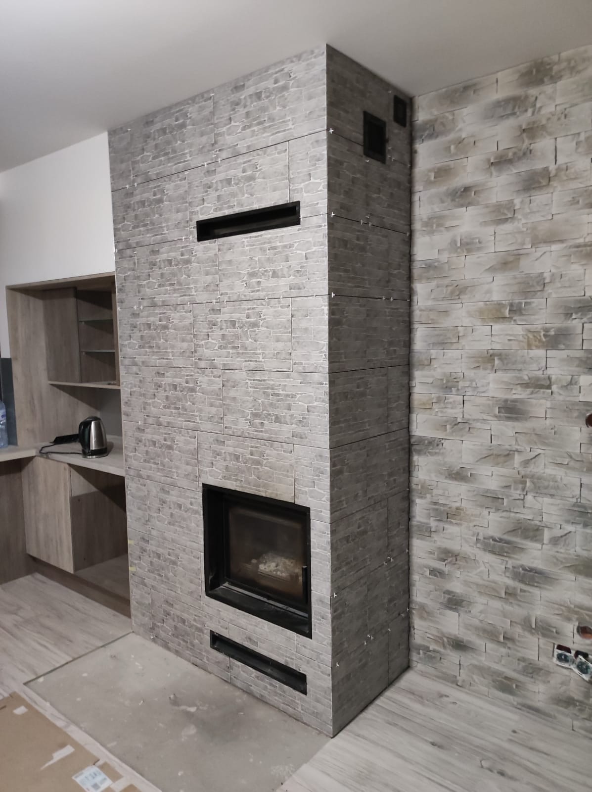 Obudowa kominka wykończona szarą okładziną imitującą kamień, z widocznymi fugami i czarnym wkładem kominkowym. Obok fragment ściany wykończony takim samym materiałem, w tle zabudowa kuchenna...