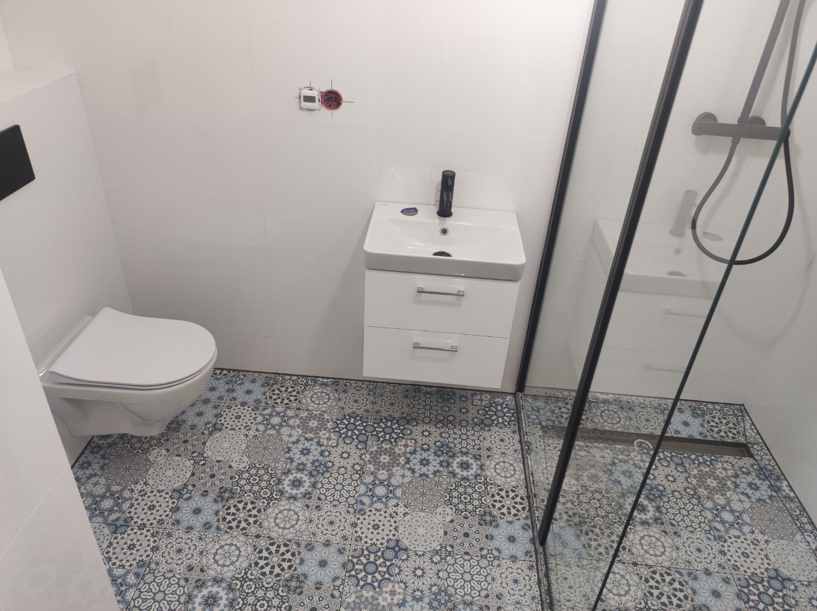 Nowoczesna łazienka z białą armaturą, wzorzystą podłogą w odcieniach szarości i błękitu oraz kabiną prysznicową z czarnymi profilami.