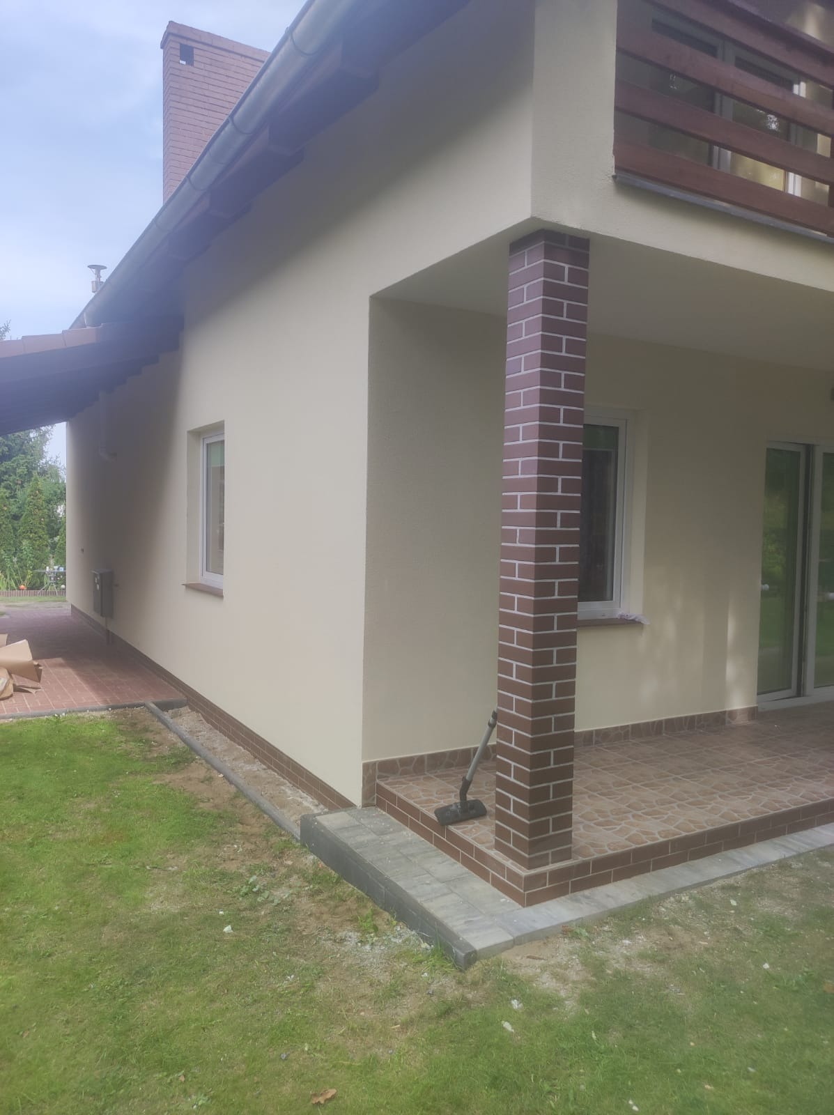 Elewacja domu jednorodzinnego w kolorze beżowym, z kolumną wykończoną klinkierem, widok z zewnątrz na taras z brązową terakotą, trawnik i fragment ogrodu.