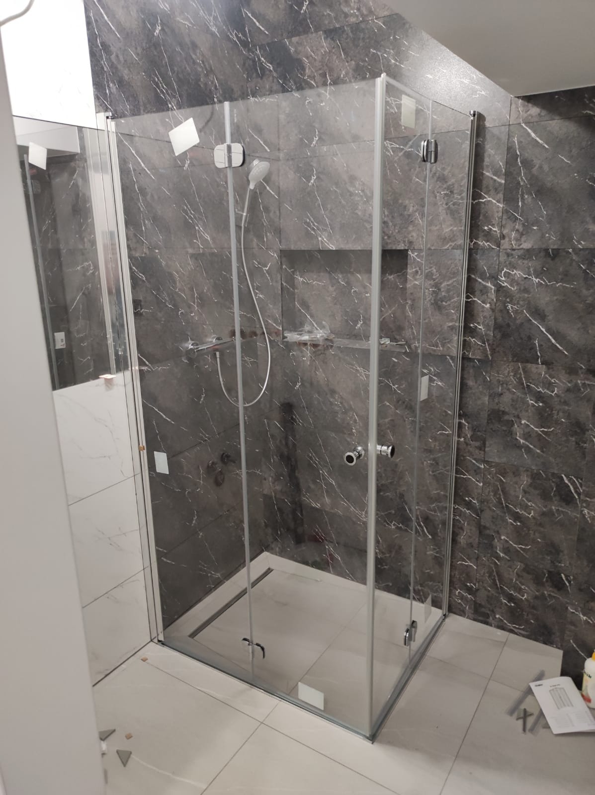 Kabina prysznicowa z transparentnego szkła, zamontowana w łazience wykończonej płytkami imitującymi ciemny marmur z białymi żyłami, z widoczną wnęką w ścianie oraz elementami armatury.