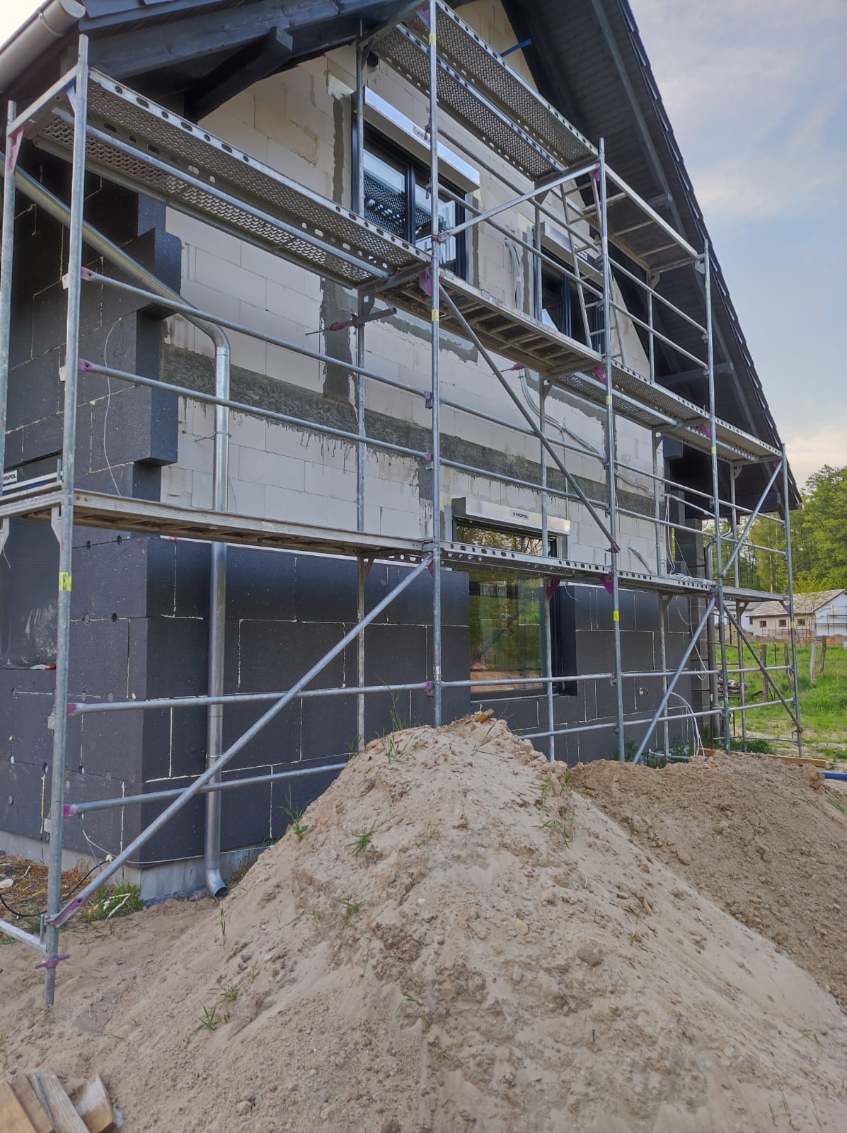 Ocieplanie domu styropianem grafitowym i białym bloczkiem, widoczne rusztowanie i hałda piasku.