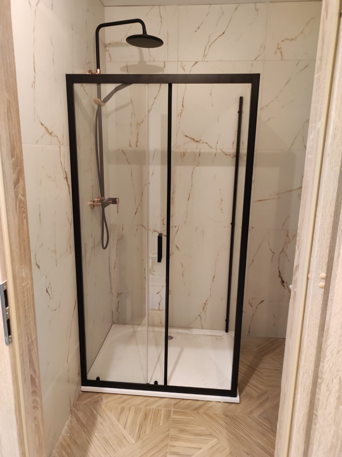 Nowoczesna kabina prysznicowa z czarną ramą i armaturą w kolorze miedzi, zamontowana w łazience wykończonej płytkami imitującymi marmur i drewnopodobną podłogą.