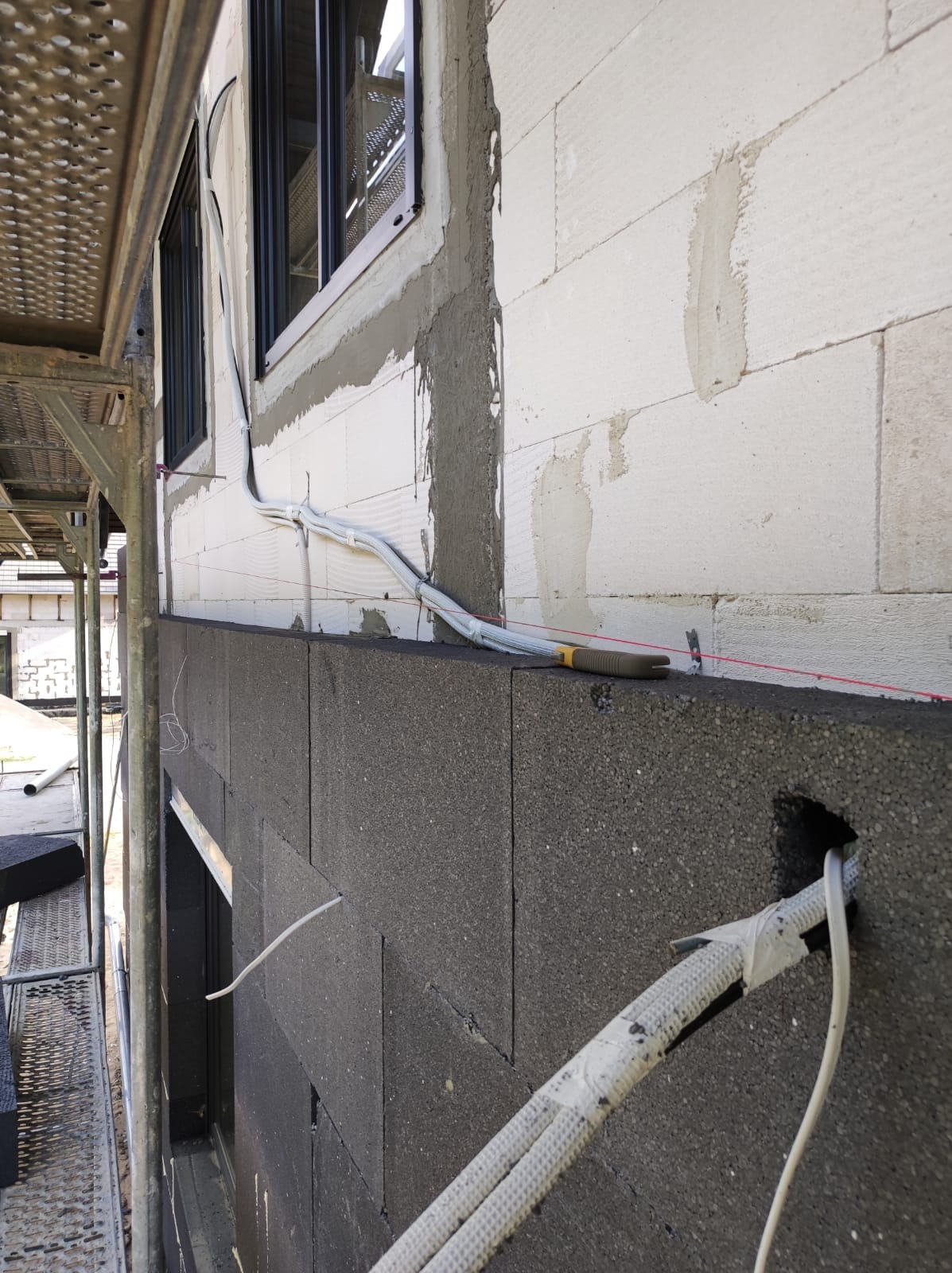 Ocieplenie budynku z szarym styropianem, widoczne kable elektryczne wychodzące z otworu w styropianie, poziomica na krawędzi styropianu i rusztowanie z boku.