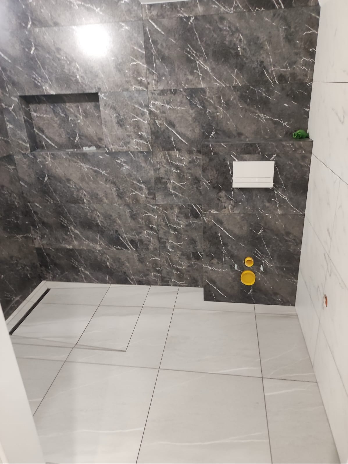 Wykończona łazienka z szarymi płytkami imitującymi marmur na ścianach, wnęką w ścianie, białymi płytkami podłogowymi i podtynkową spłuczką WC. Widoczne żółte zaślepki instalacji wodnej.
