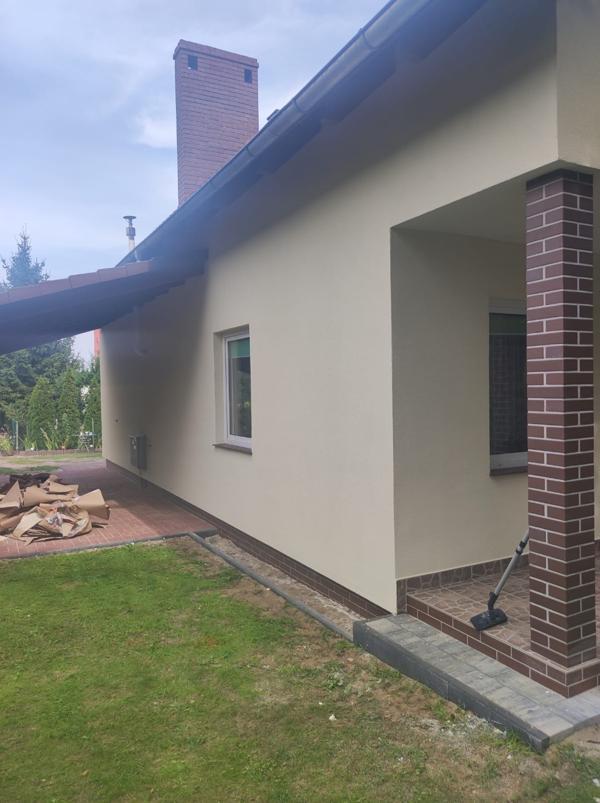 Elewacja domu jednorodzinnego po nałożeniu tynku strukturalnego w odcieniach beżu, z widocznym fragmentem ceglanego komina i filaru werandy, na trawniku leżą sterty papierowych worków po...