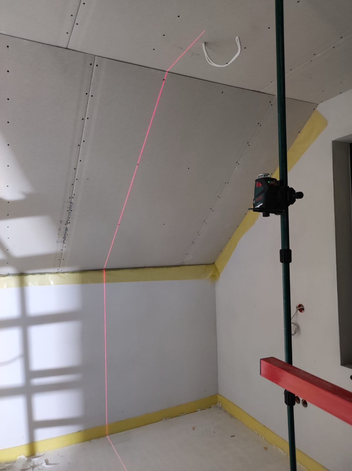 Montaż płyty karton gips na poddaszu z użyciem poziomicy laserowej, widoczne zabezpieczenia folią i taśmą malarską.