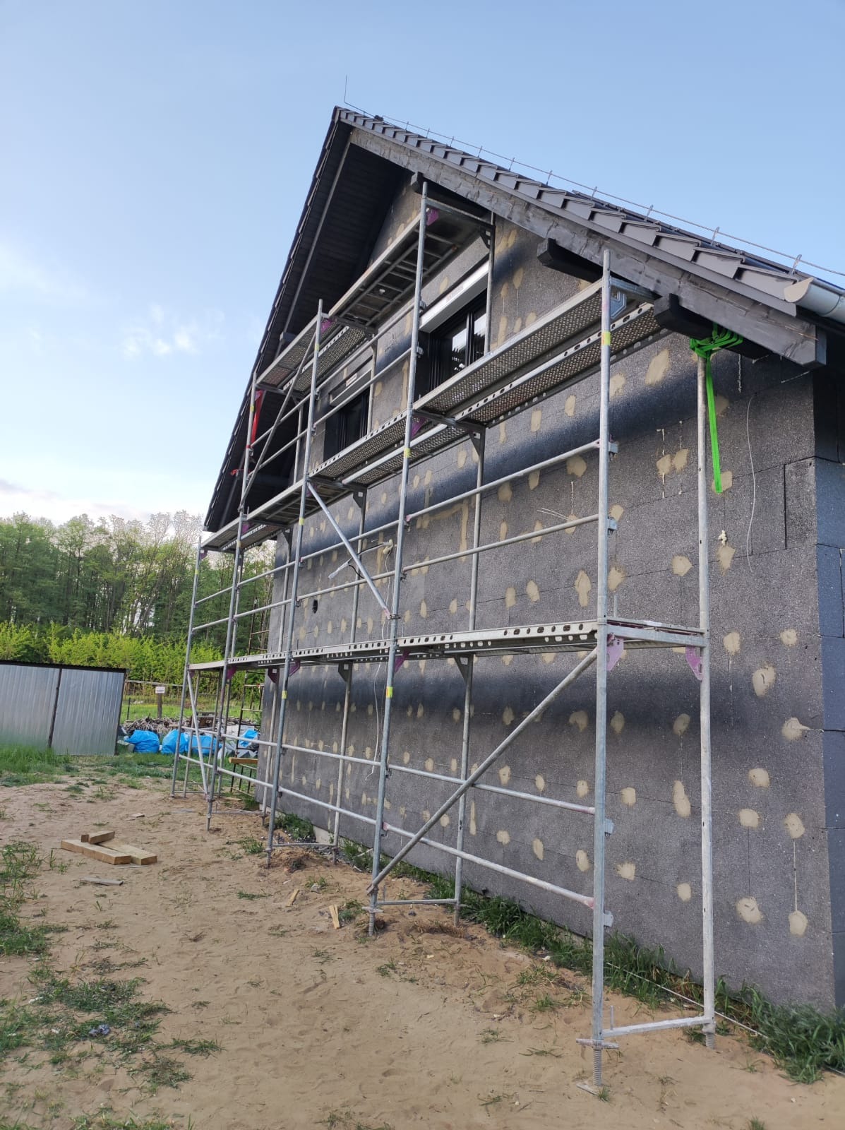 Dom w trakcie ocieplania, widoczne rusztowanie, styropian z kołkami, szara elewacja, przygotowanie do dalszych prac wykończeniowych.