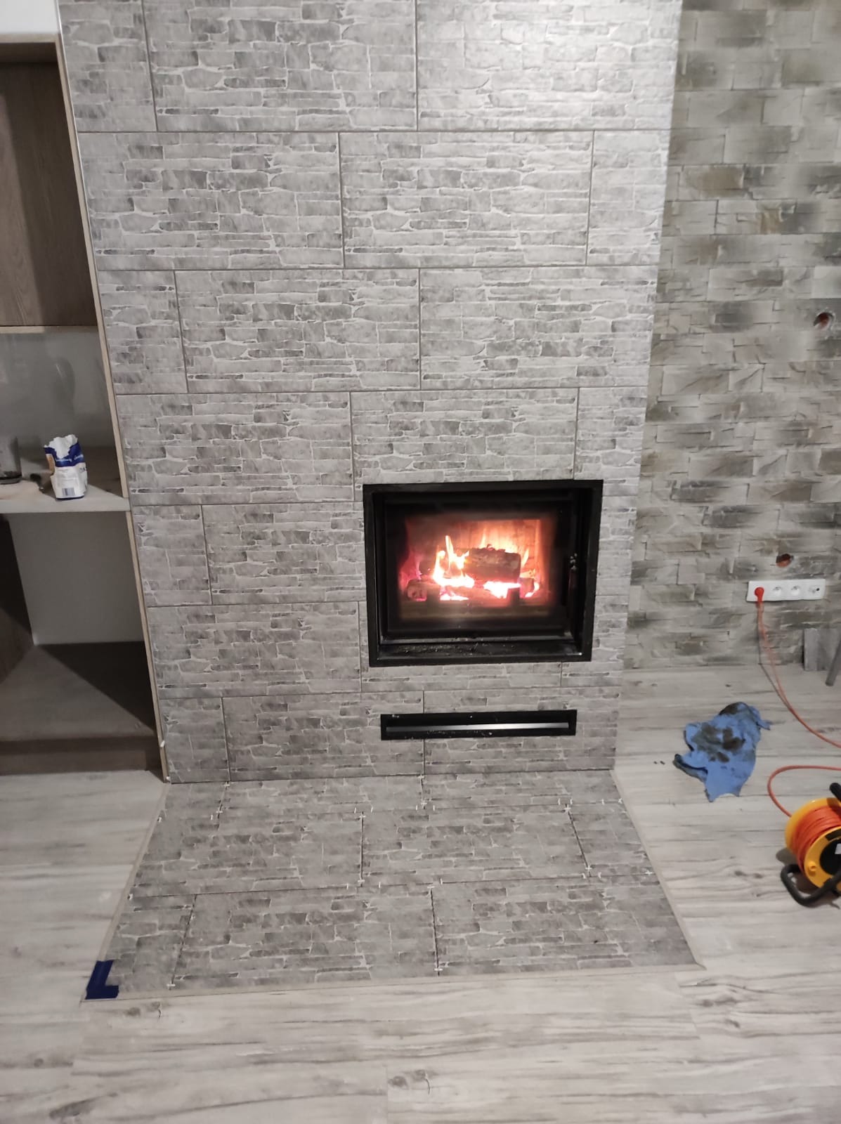 Obudowa kominka wykończona szarą, kamienną okładziną, z widocznym paleniskiem i akcesoriami do prac wykończeniowych w tle.