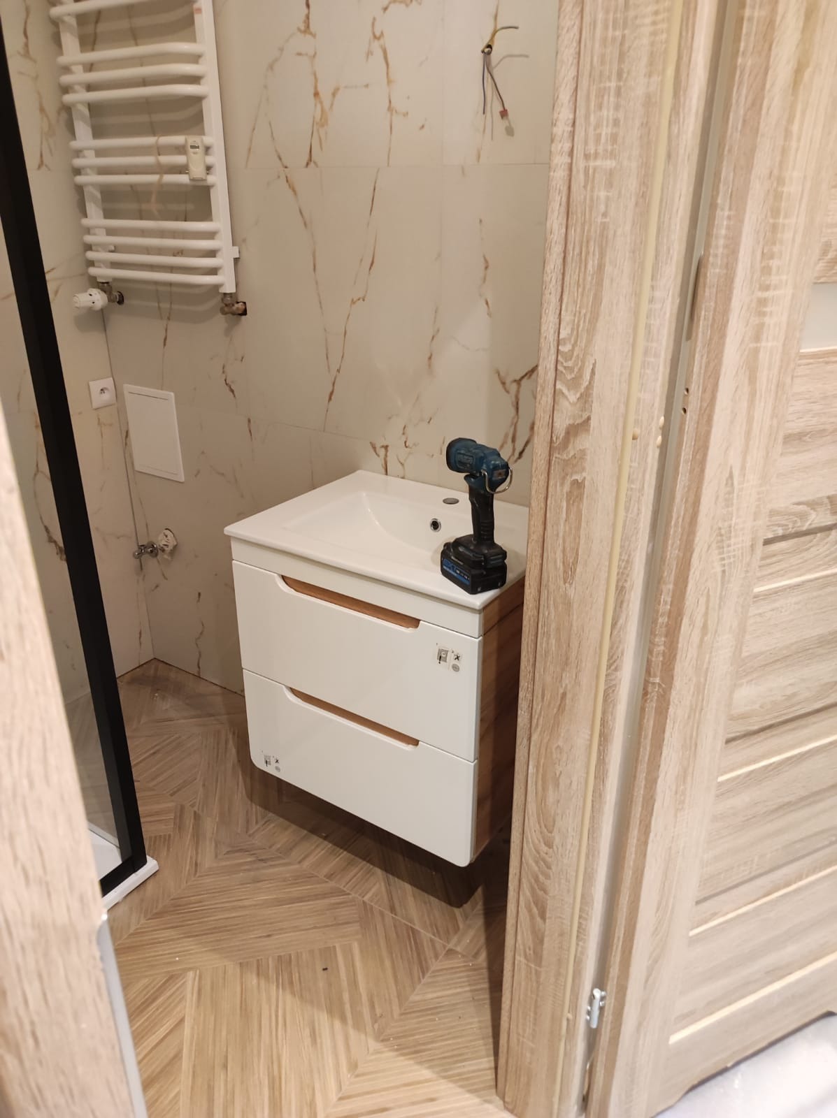 Biała szafka pod umywalkę z dwiema szufladami w łazience z marmurowymi płytkami, wiertarka akumulatorowa leży na umywalce, widoczny grzejnik drabinkowy i fragment kabiny prysznicowej.