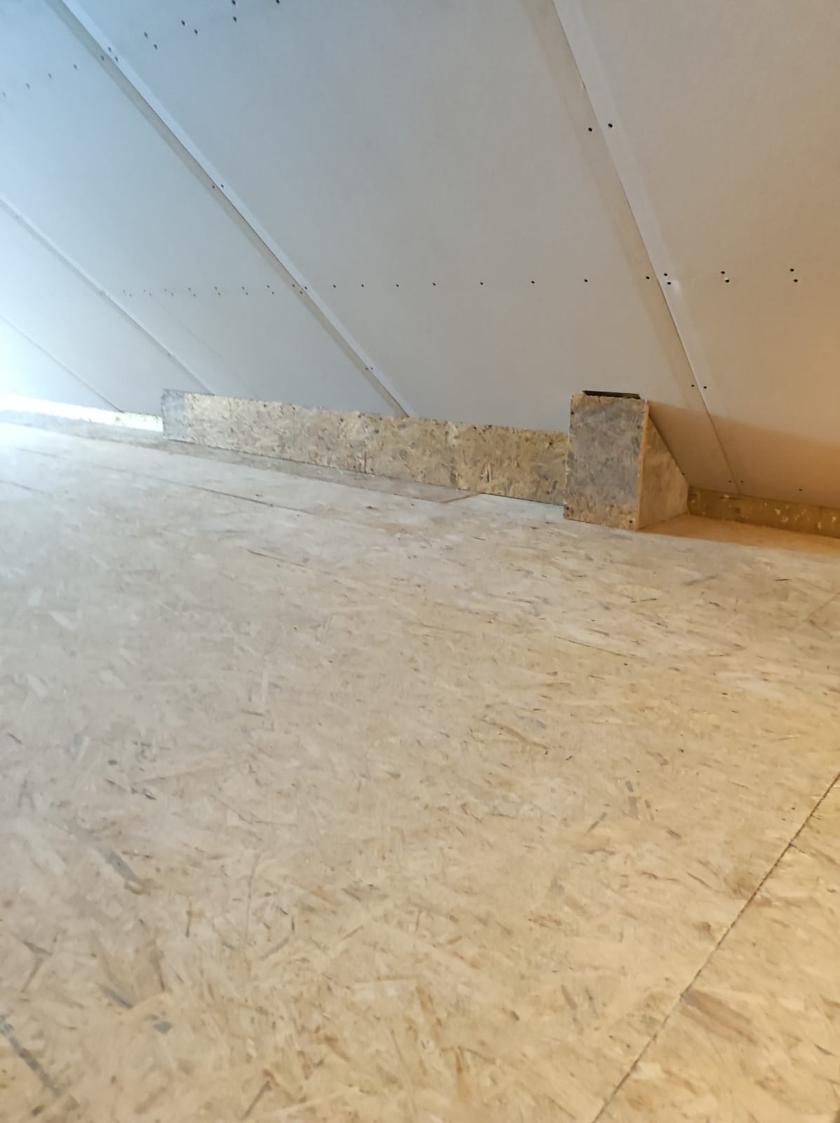 Skośne poddasze z płytami OSB na podłodze i zamontowanymi płytami gipsowo-kartonowymi na skosach dachu, widoczne fragmenty drewnianej konstrukcji wzmacniającej.