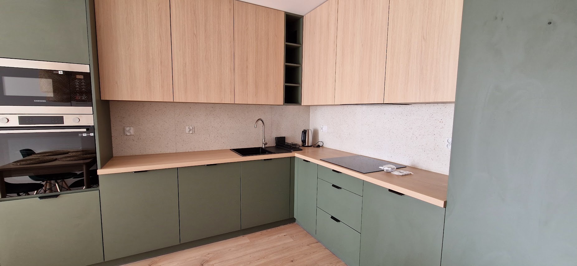 Nowoczesna kuchnia z zielonymi szafkami dolnymi i drewnianymi górnymi, czarnym zlewem i płytą indukcyjną, wykończona blatem w kolorze jasnego drewna i mozaiką na ścianie.