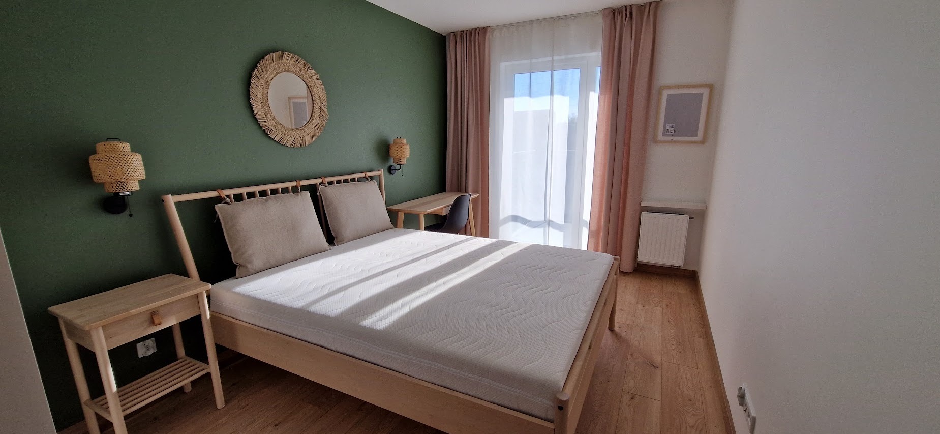 Jasna sypialnia z łóżkiem z drewnianą ramą, zieloną ścianą z okrągłym lustrem z rafii i rattanowymi lampami ściennymi, drewnianą podłogą i biurkiem przy oknie.