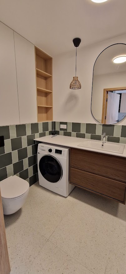 Nowoczesna łazienka z pralką zabudowaną pod blatem, zielono-szara mozaika na ścianie, wisząca szafka z drewnianymi frontami, owalne lustro z czarną ramą i lampa z plecionki.