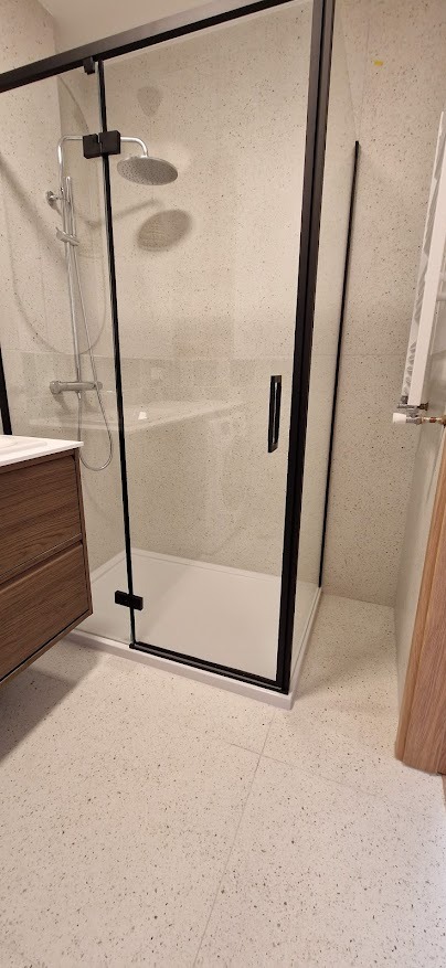 Nowoczesna kabina prysznicowa z czarną ramą, deszczownicą i drewnianą szafką umywalkową na tle jasnych płytek z drobnymi plamkami.