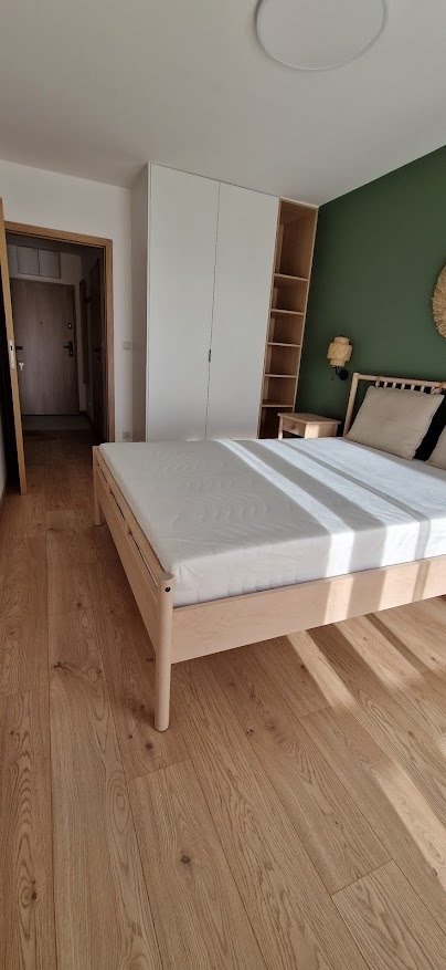 Jasne drewniane panele podłogowe w sypialni z łóżkiem z jasnego drewna, białą szafą i zieloną ścianą z dekoracją, widok na korytarz.