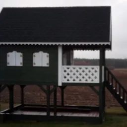 Mała, drewniana konstrukcja przypominająca domek na podwyższeniu z zadaszeniem, schodami i ozdobnymi okiennicami, ustawiona na tle zaoranego pola i lasu w pochmurny dzień.