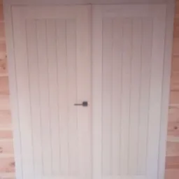 Dwuskrzydłowe, jasne drzwi wewnętrzne z pionowymi panelami i czarną, prostokątną klamką na tle boazerii.