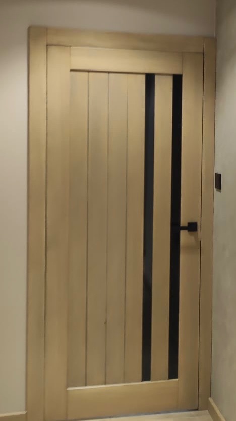 Beżowe drzwi wewnętrzne z pionowymi panelami i dwoma wąskimi, czarnymi wstawkami, z czarną klamką.