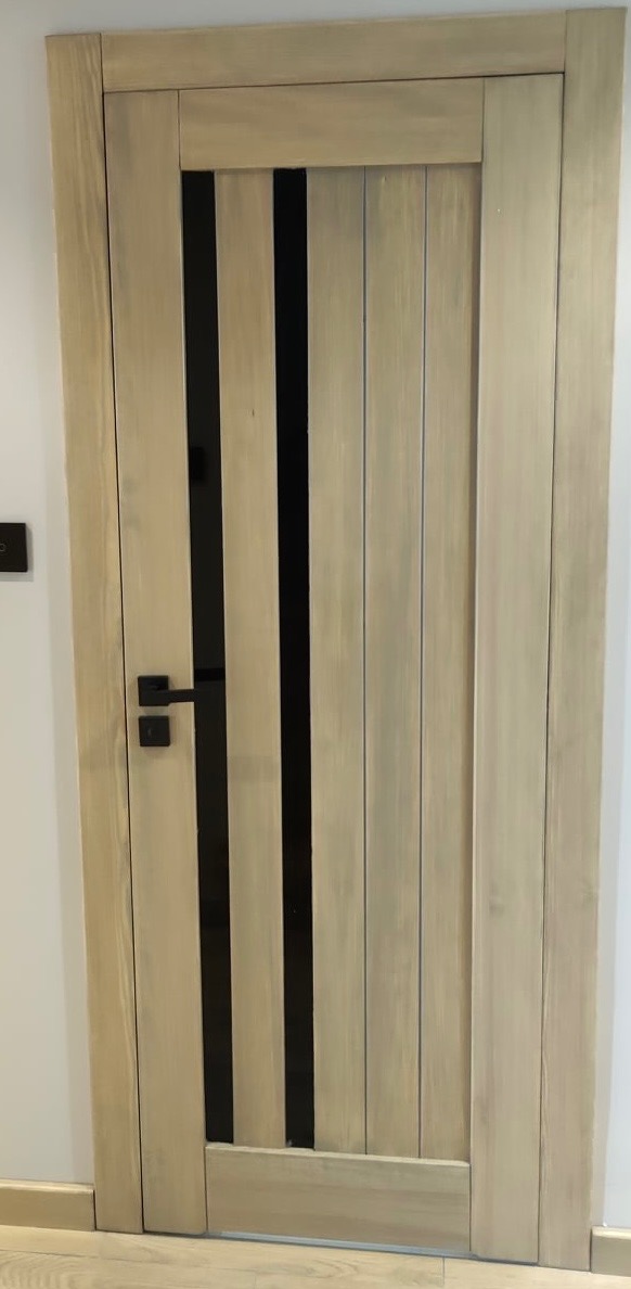 Drewniane drzwi wewnętrzne w odcieniach beżu z czarnymi, pionowymi wstawkami i czarną klamką, widoczne na tle białej ściany i jasnej podłogi.