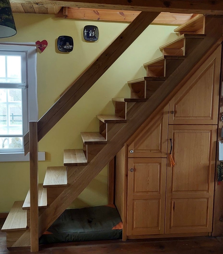 Drewniane schody z jasnego drewna, wkomponowane w zabudowę szafek, pod oknem z dekoracjami na ścianie w ciepłym odcieniu żółci.