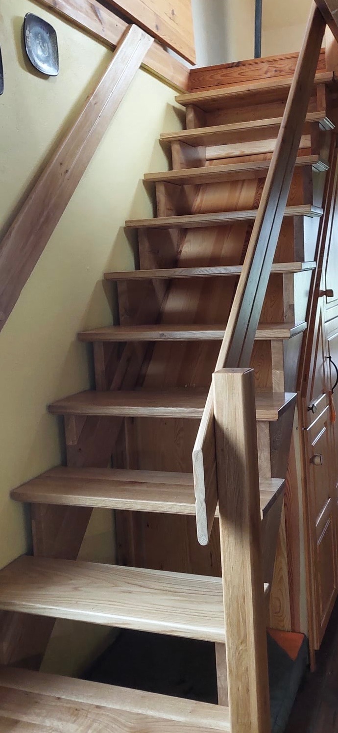 Drewniane schody wewnątrz domu, z jasnego drewna, z prostą balustradą i widoczną fakturą drewna, na tle żółtej ściany i drewnianej szafy.