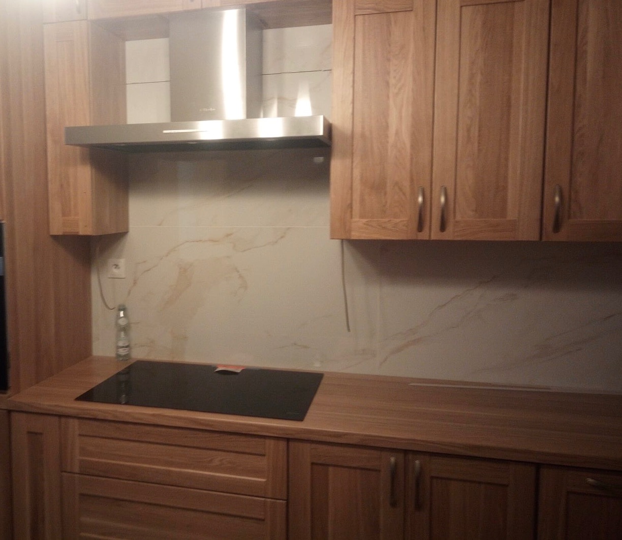 Fragment drewnianej zabudowy kuchennej z płyta indukcyjną i okapem, marmurkowe płytki na ścianie.