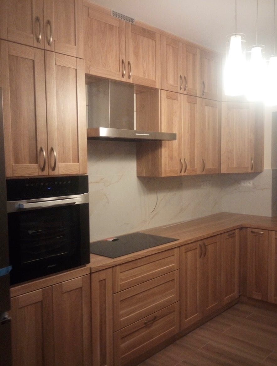 Jasna kuchnia z drewnianymi frontami, czarnym piekarnikiem i płytą grzewczą, oświetlona wiszącymi lampami z mlecznego szkła.