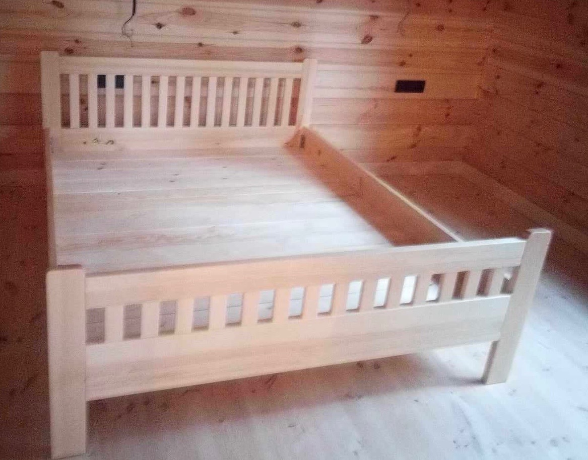 Surowe, niedokończone łóżko z jasnego drewna w prostym stylu, widoczne niedoskonałości obróbki, na tle drewnianej ściany i podłogi.