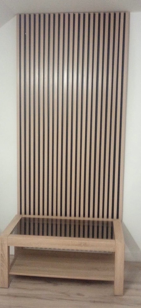 Nowoczesny panel ścienny z jasnego drewna z czarnymi wstawkami, tworzący dekoracyjne tło dla niskiego stolika z blatem z czarnego szkła i dodatkową półką.